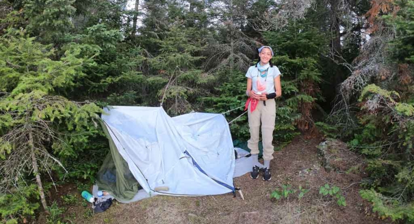 an outward bound student stands beside their tarp shelter in a wooded area 